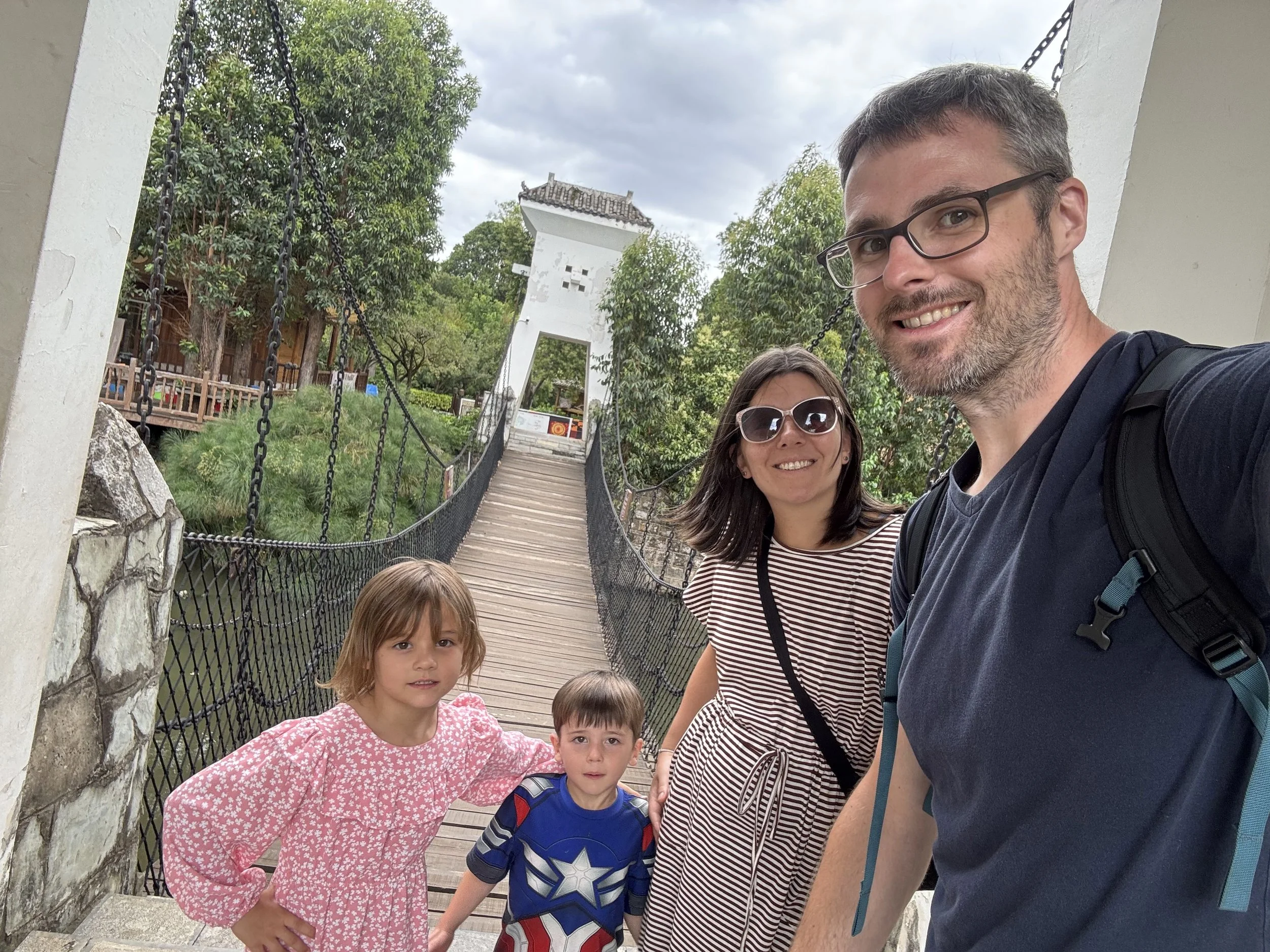 A group of four people, including a man, a woman, and two children, standing on a wooden bridge with chains on either side. Behind them is a white archway and lush green trees. The sky is cloudy.