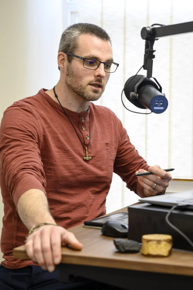A man with glasses and a beard sitting at a desk with a microphone, holding a phone, in a well-lit room.