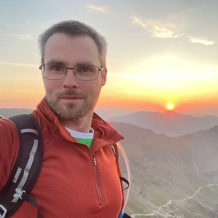 A man with glasses and a beard taking a selfie during sunrise or sunset in a mountainous area. He is wearing a red jacket and carrying a backpack.
