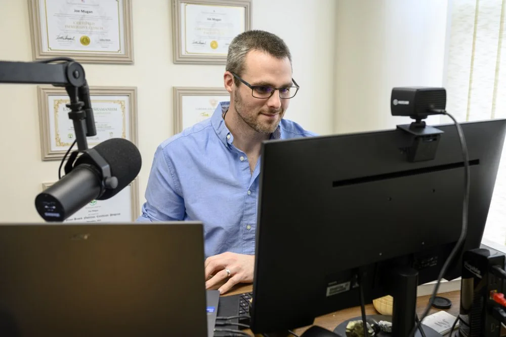 A man with glasses and a blue shirt sitting at a desk, working on a computer in a home office, with framed certificates on the wall behind him.