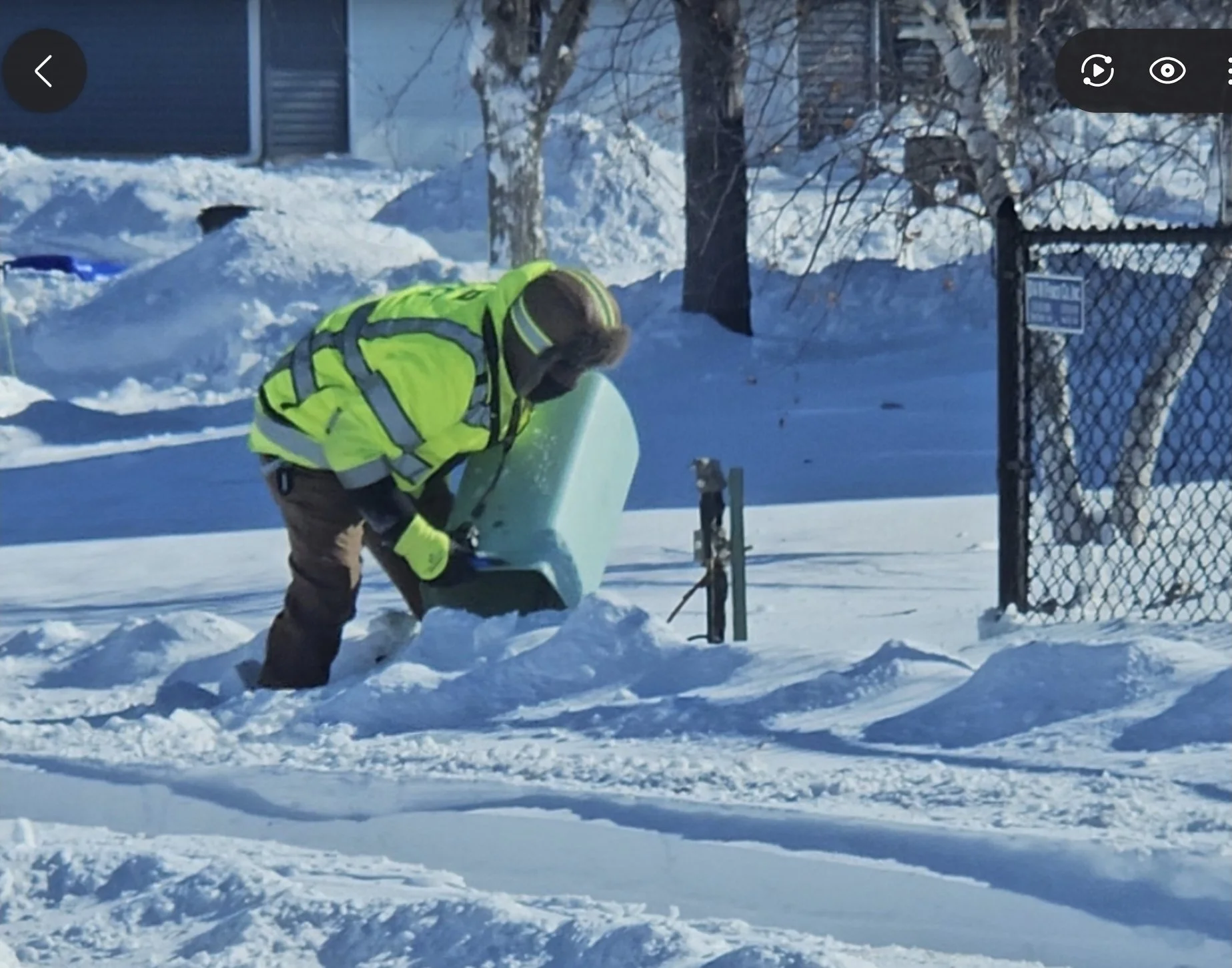 A person in a bright yellow jacket and brown pants is shoveling snow in a backyard, with snow-covered ground, trees, a fence, and a slide visible in the background.