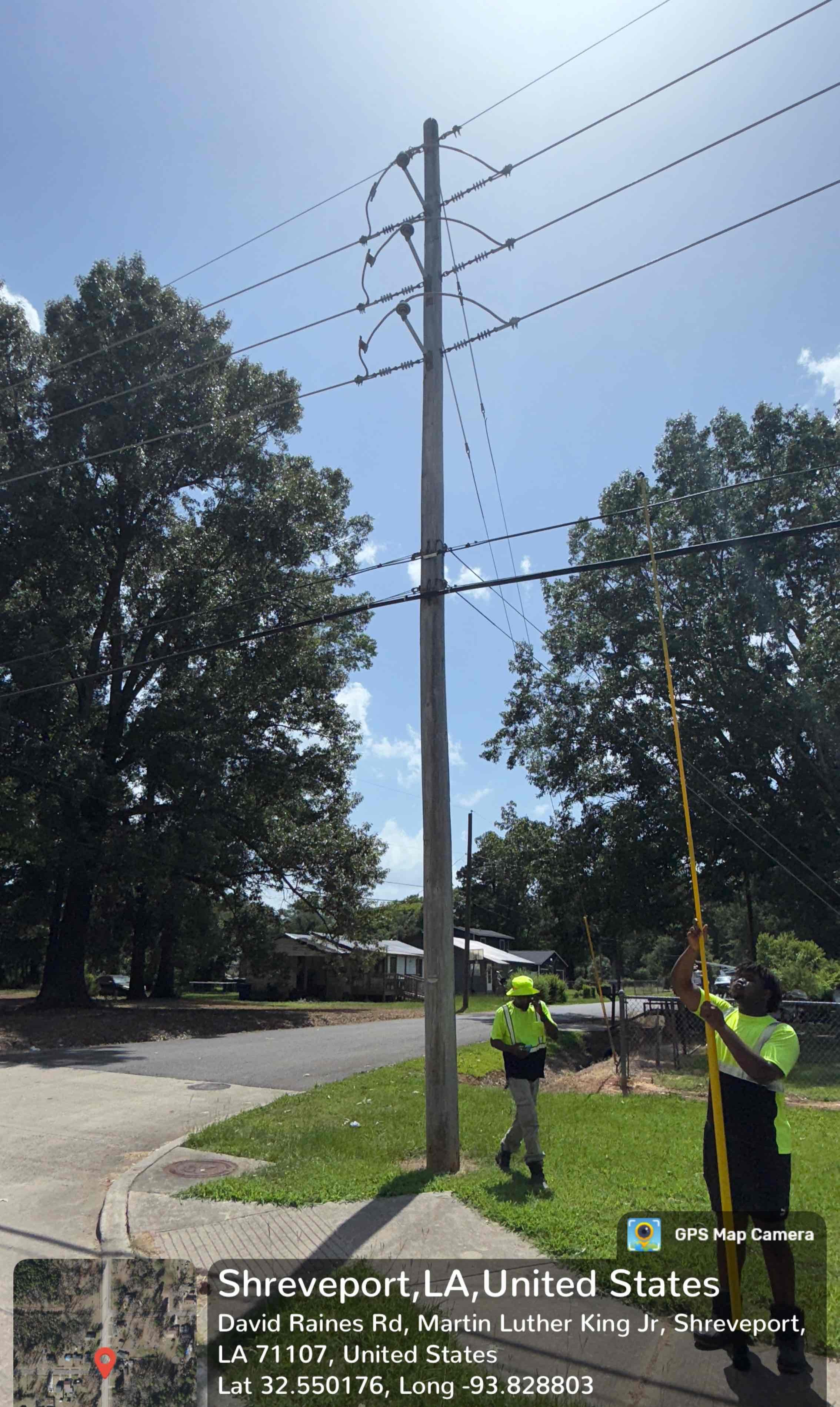 Utility workers in safety vests and helmets repair or inspect a tall wooden utility pole with electrical wires on a sunny day in a residential neighborhood in Shreveport, Louisiana. Trees and houses are visible in the background.