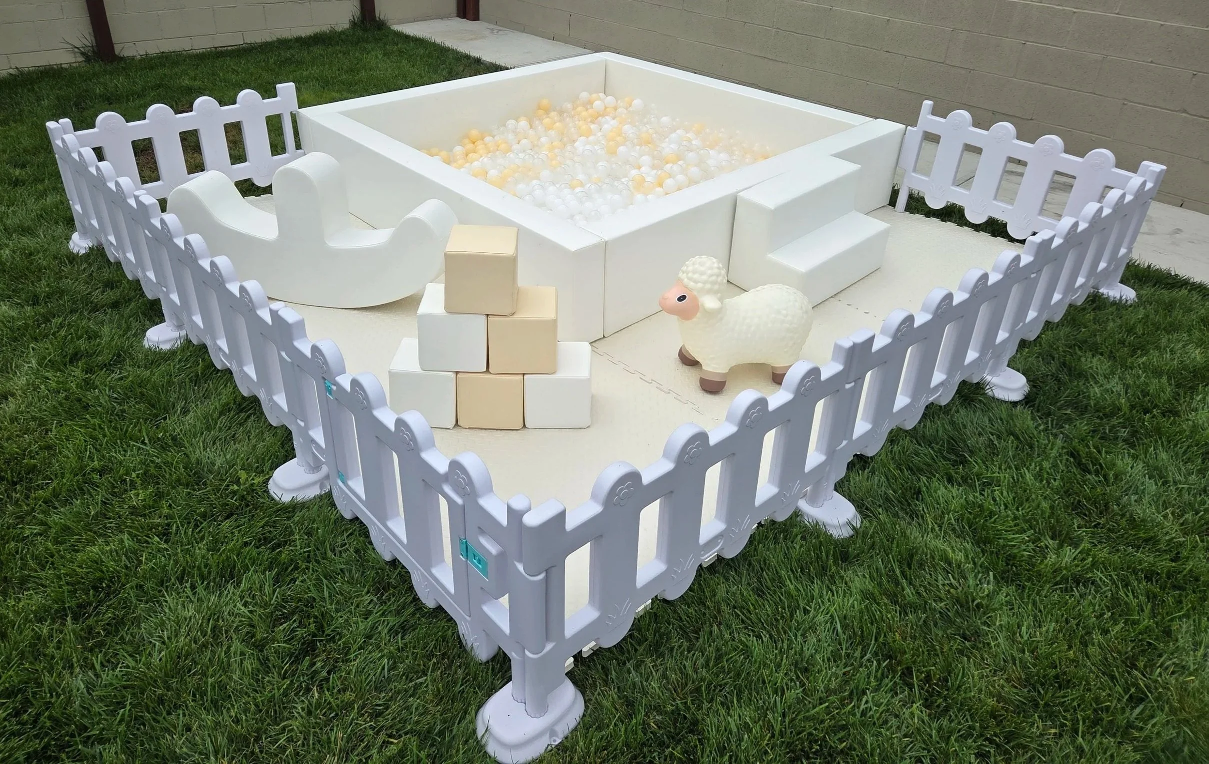 A white ball pit filled with white, clear, and beige balls, surrounded by a white safety fence. There is a small white toy sheep and cream-colored blocks near the entrance. The setup is outdoors on grass with a brick and wall background.