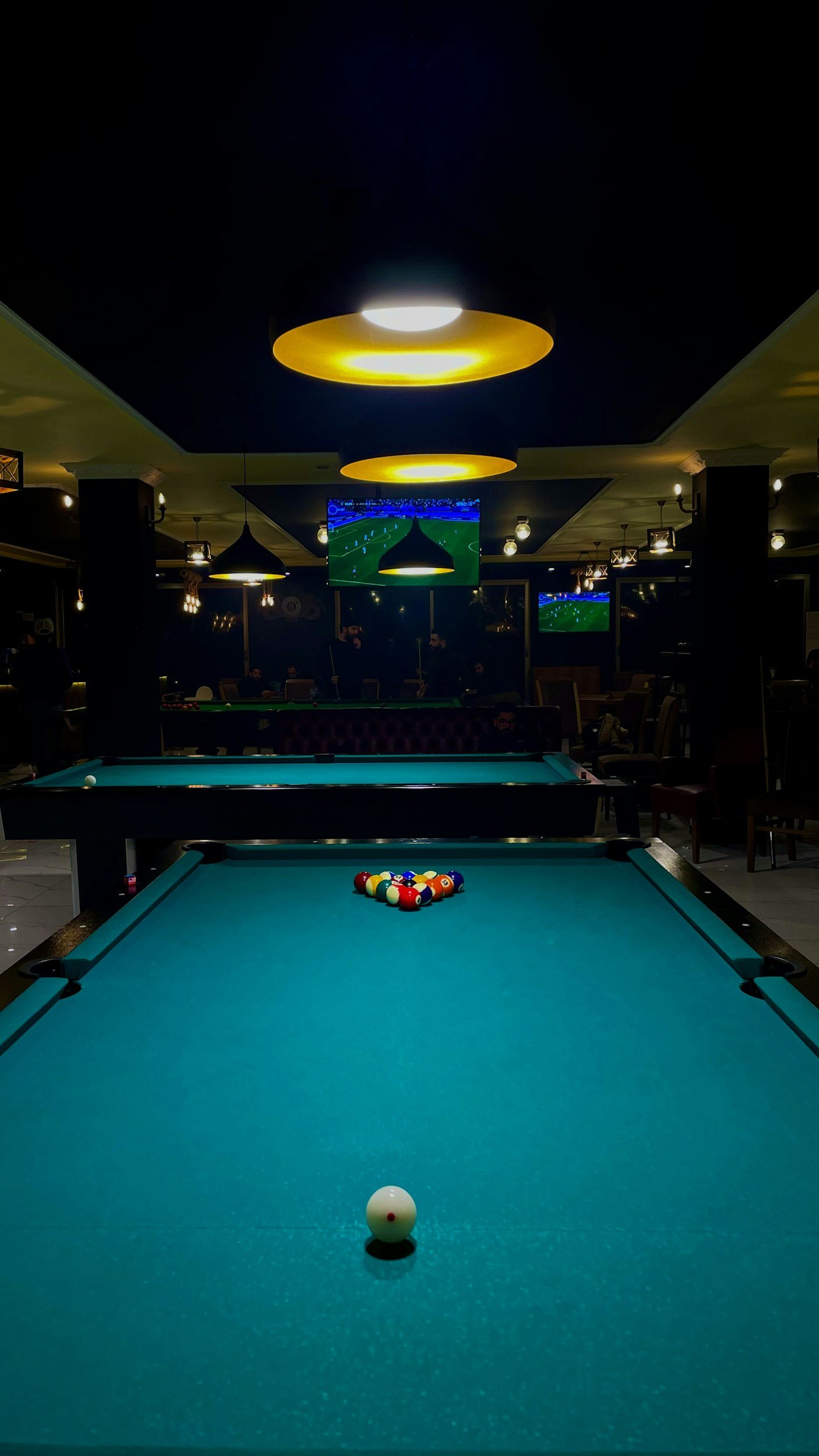 Billiard table with balls racked, and a white cue ball in the foreground, in a dimly lit bar or pool hall with multiple television screens showing sports.