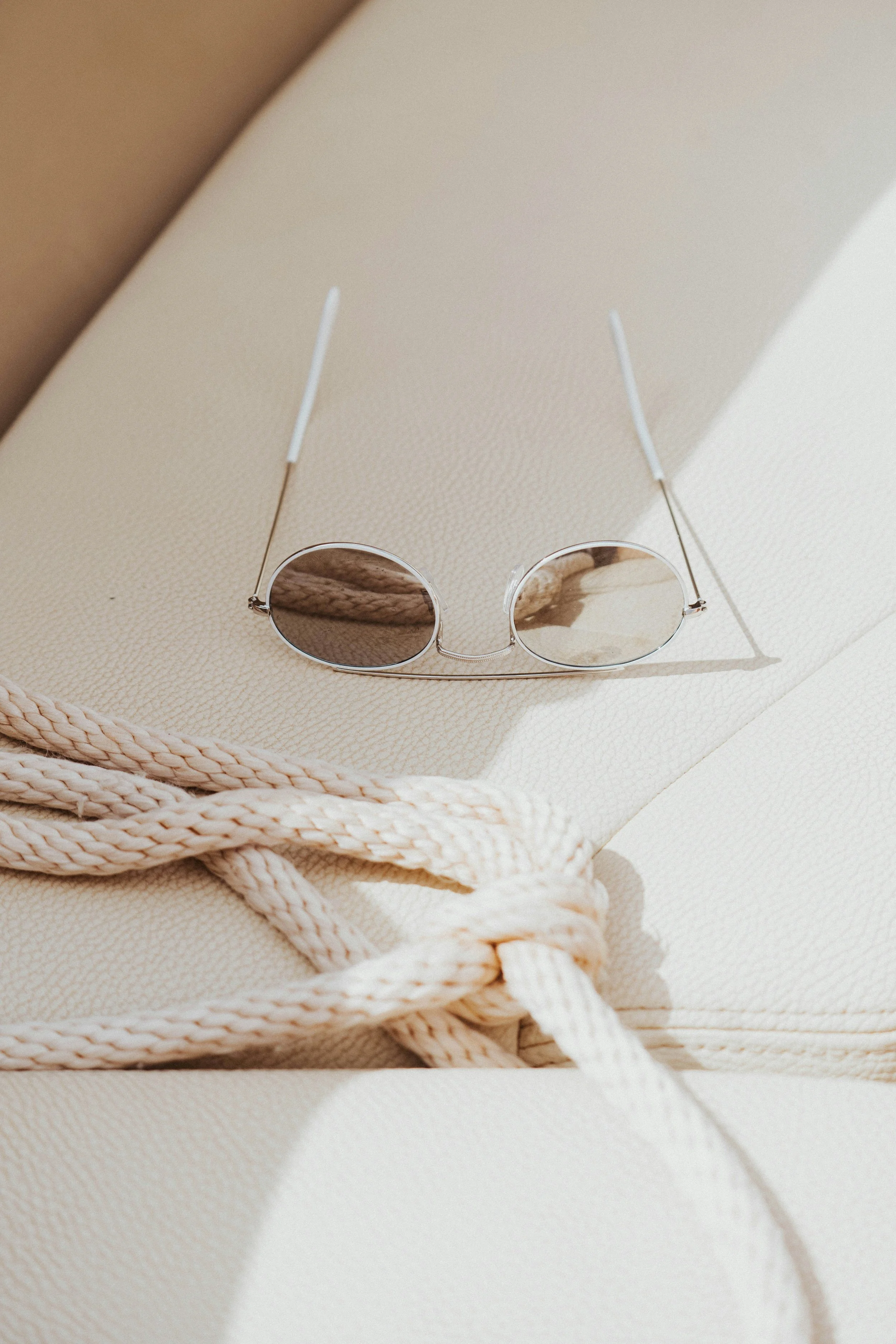 Round sunglasses with thin metal frames placed on light-colored leather, next to a knotted rope.