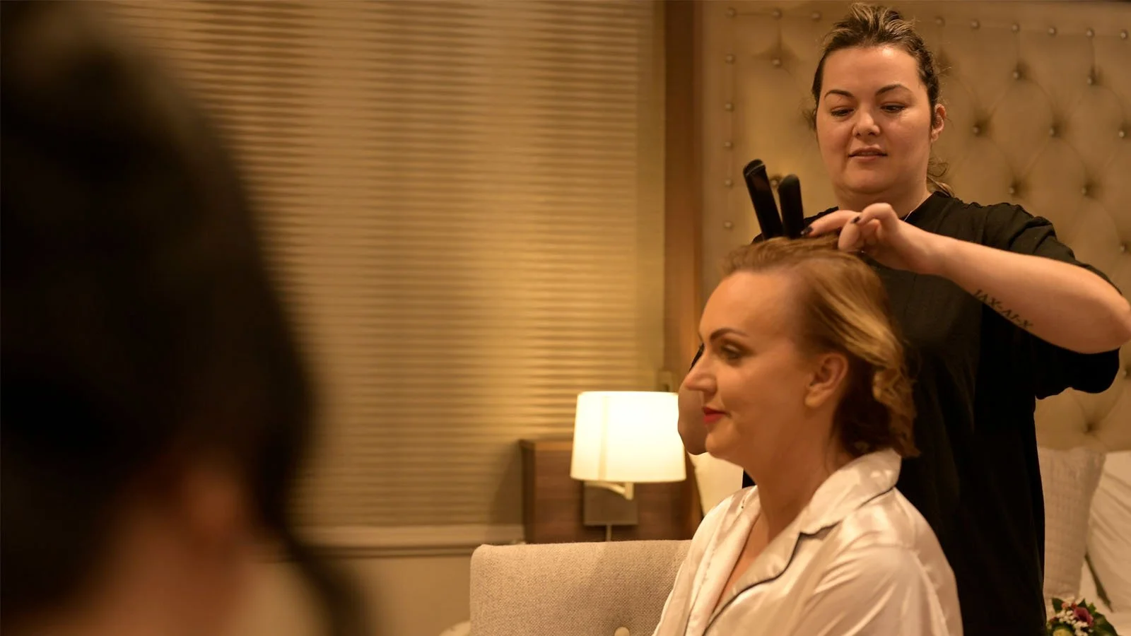 The bride is sitting in the bedroom while the hairstylist fixes her hair.