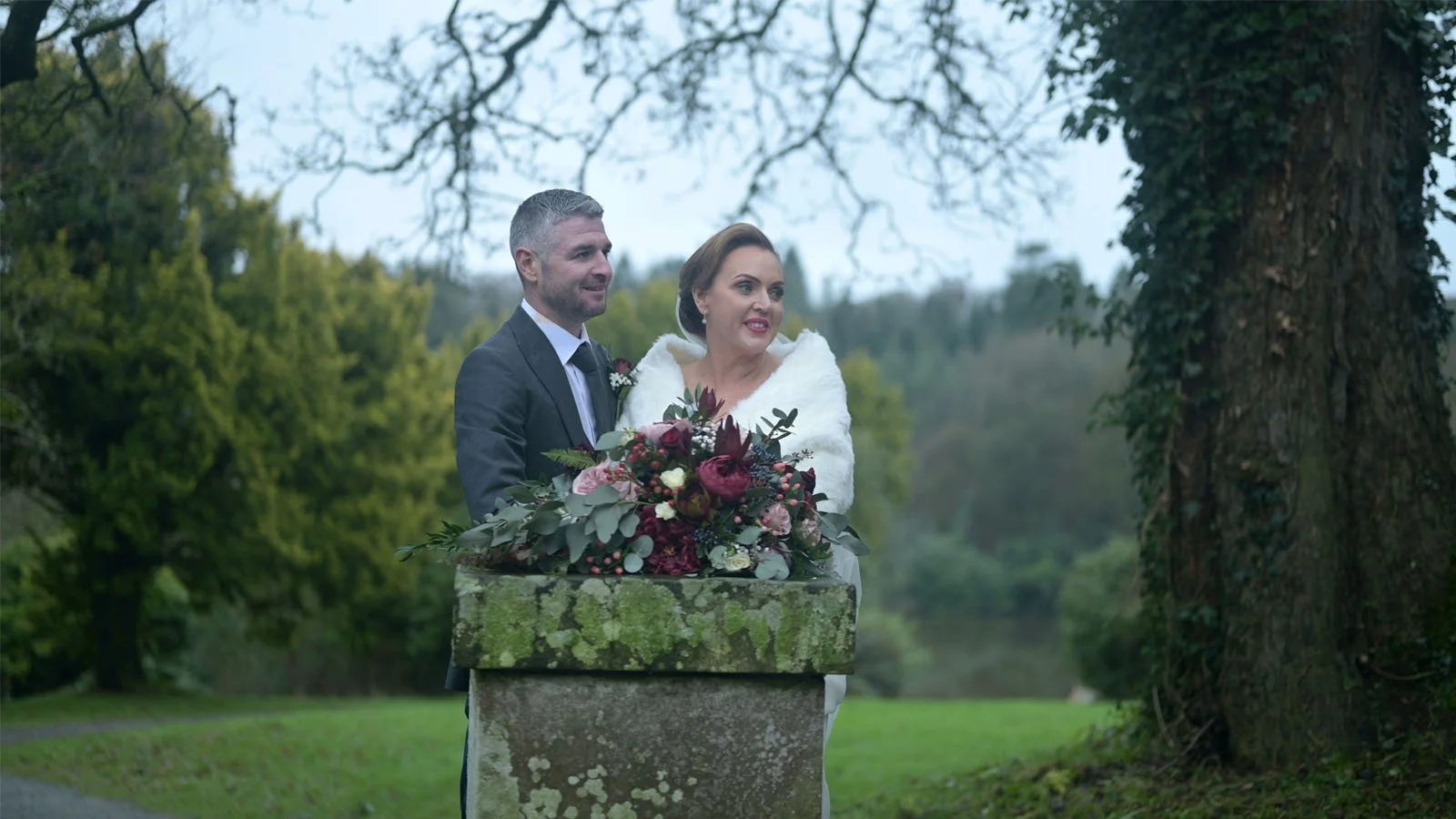  A married couple standing beside an old stone pillar with plenty of trees behind and around them. 
