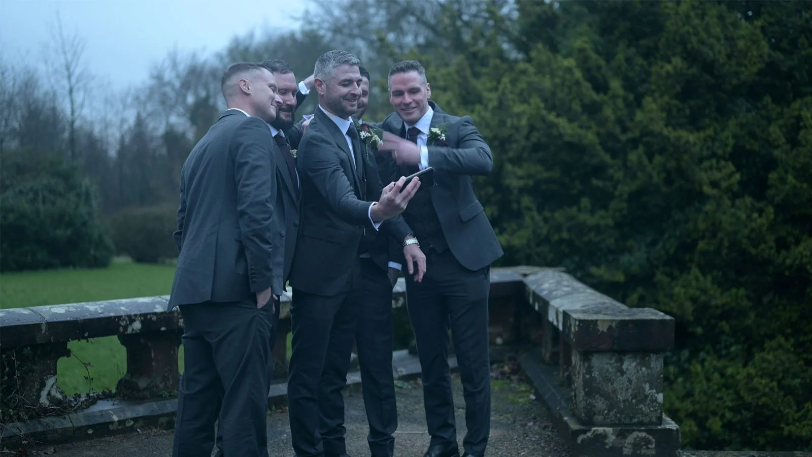  Groom with groomsmen having fun beside a small old stone wall with plenty of trees around them. 