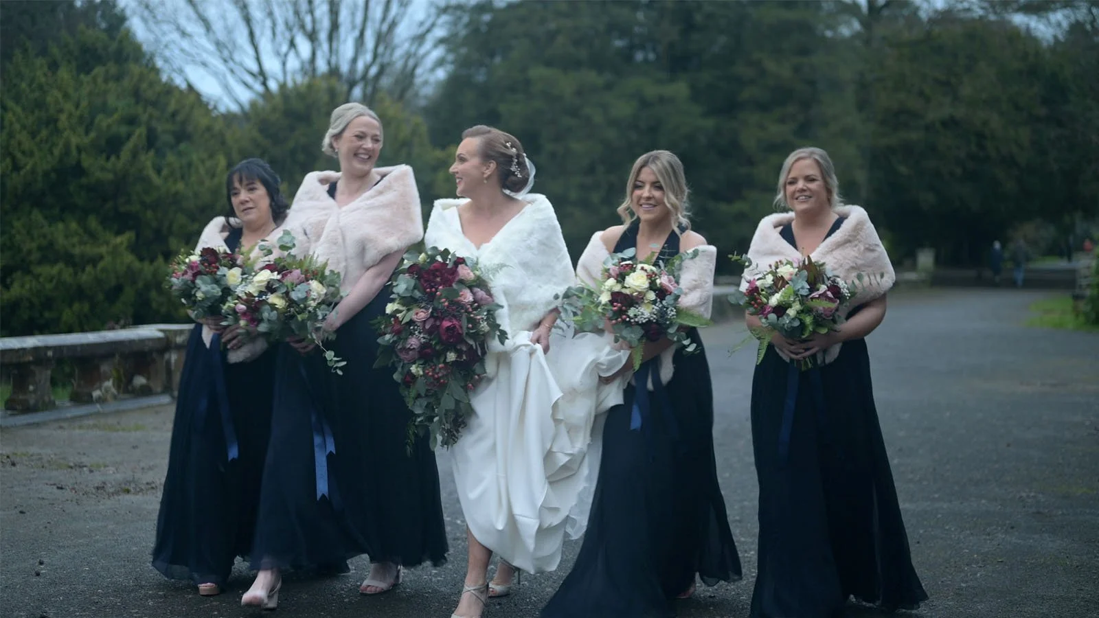  The bride and with bridesmaids walk and talk, holding bouquets along the large path. 