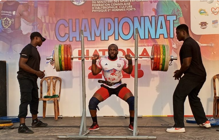 A male weightlifter in a white and red shirt and black shorts squats with a heavily loaded barbell on his shoulders. Two spotters stand nearby, one on each side, ready to assist. The background has a banner with French text and the word 'CHAMPIONNAT' in large letters.