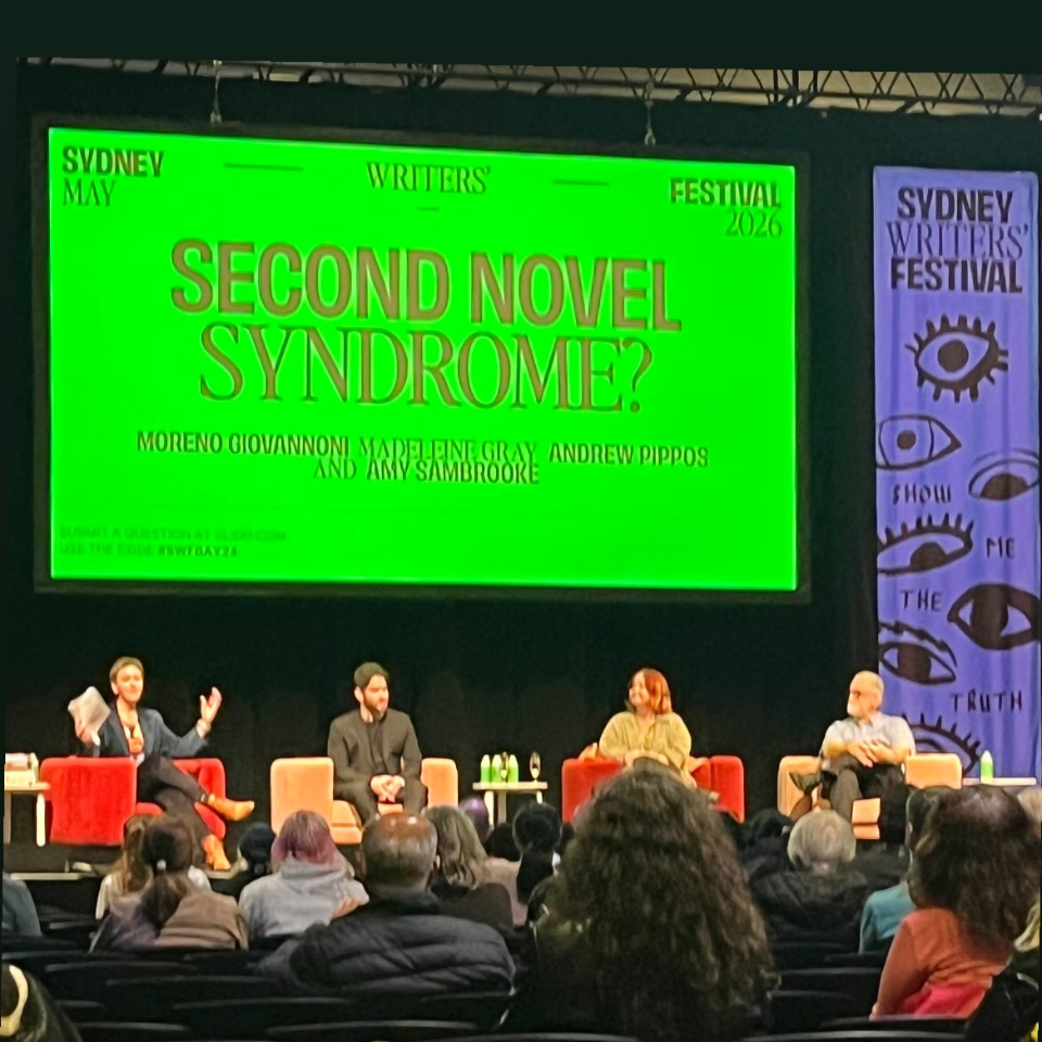 A panel discussion at the Sydney Writers' Festival in 2026 about the topic 'Second Novel Syndrome.' Four people are seated on stage, with one person speaking and gesturing. There is a large green screen behind them displaying the event details, and a