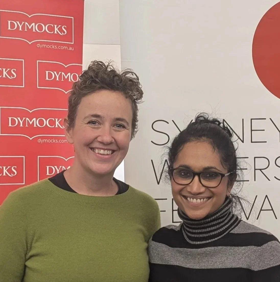 Two smiling women standing in front of event signs. The woman on the left has short curly hair and is wearing a green sweater. The woman on the right has long dark hair, glasses, and is wearing a black and gray striped turtleneck.