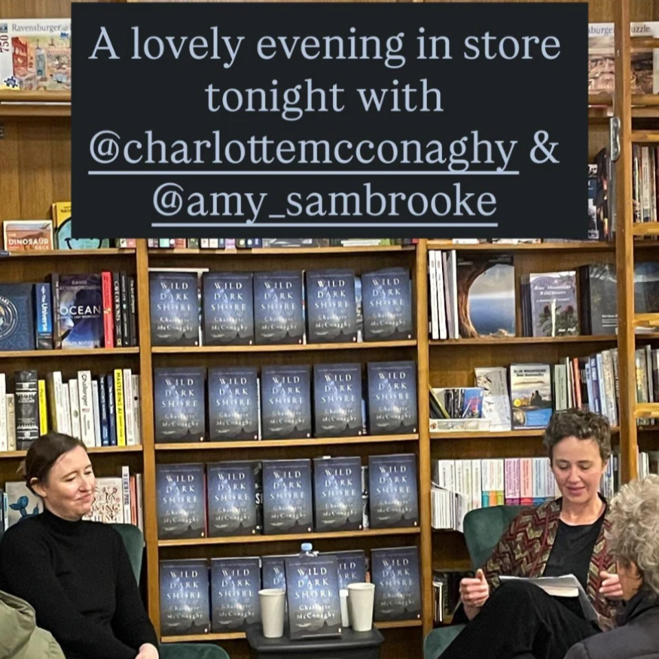 Two women sitting in chairs and a woman with glasses sitting in front of them in a bookstore, with shelves of books and a sign overhead that reads 'A lovely evening in store tonight with @charlottemcconaghy & @amy_sambrooke'.