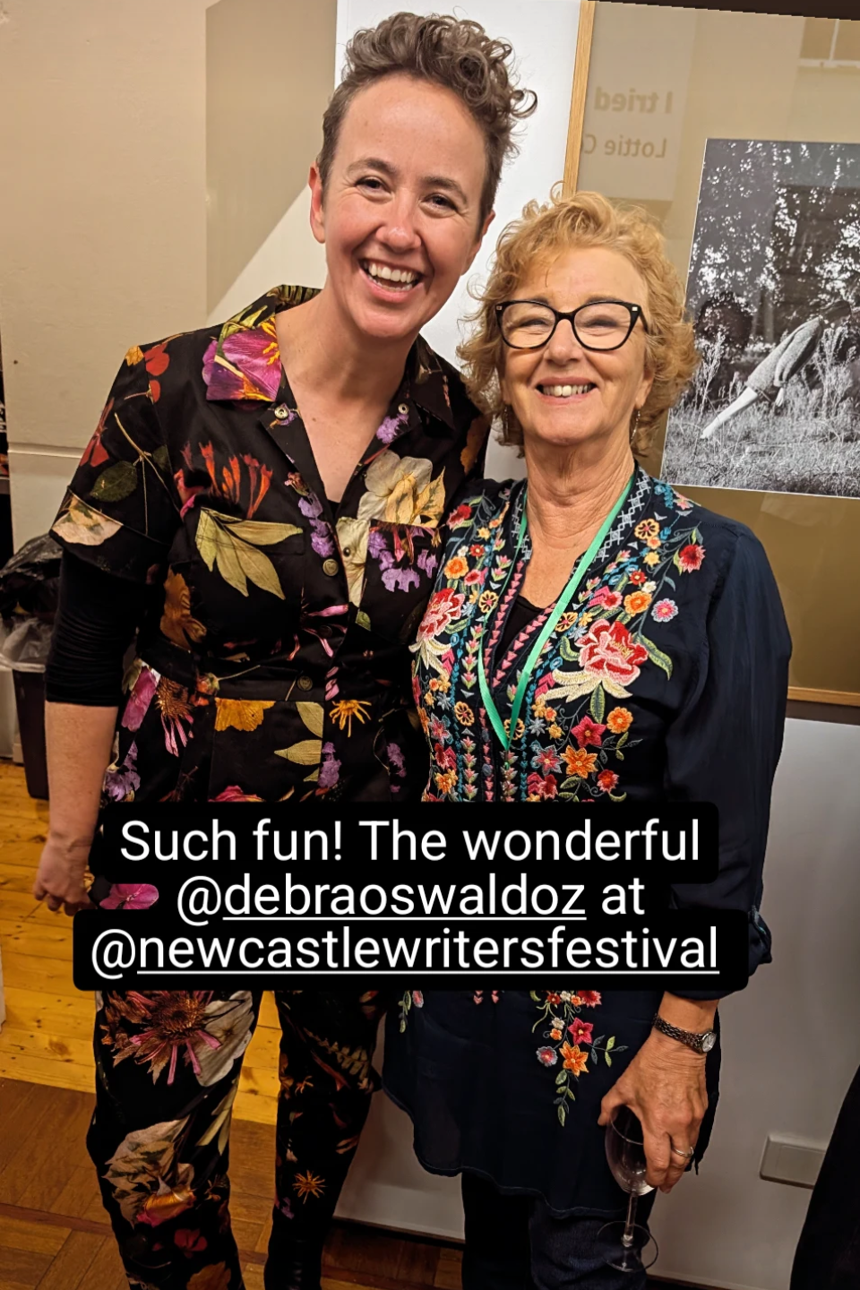 Two women smiling at a writer's festival, one wearing a floral shirt, the other in a navy embroidered blouse, standing close together in an indoor setting.