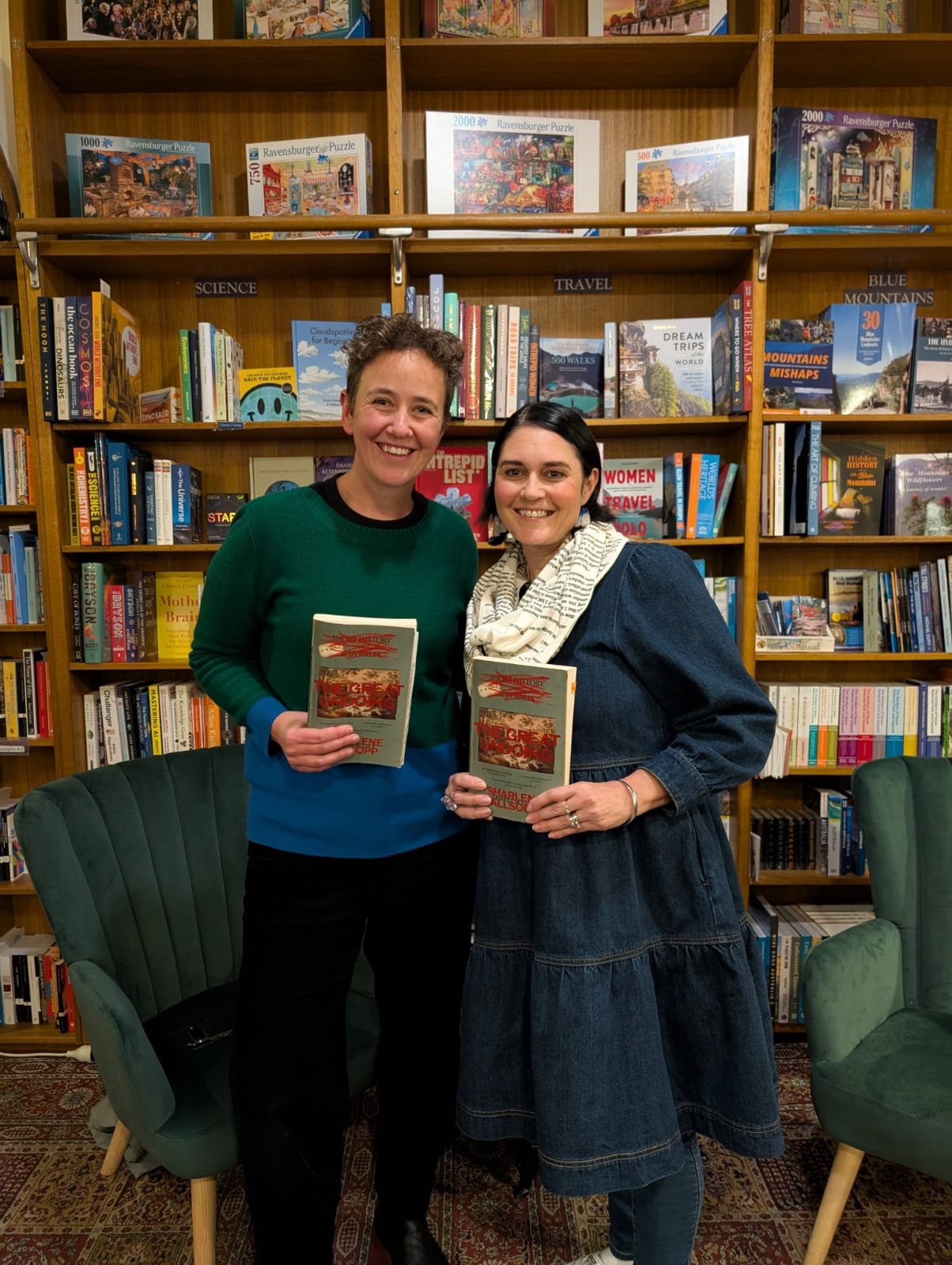 Two women standing in a bookstore, each holding a book and smiling at the camera. Behind them are wooden bookshelves filled with various books, and there are two green chairs in front of them.