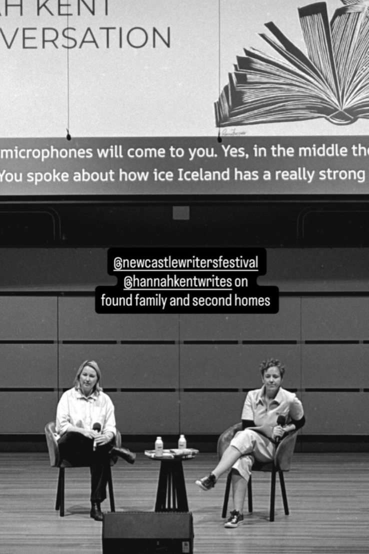 Two women sitting on stage, each holding a microphone, during a discussion at an event. There is a small table with water bottles and books between them. A large screen above displays text and an open book illustration, with social media handles superimposed.