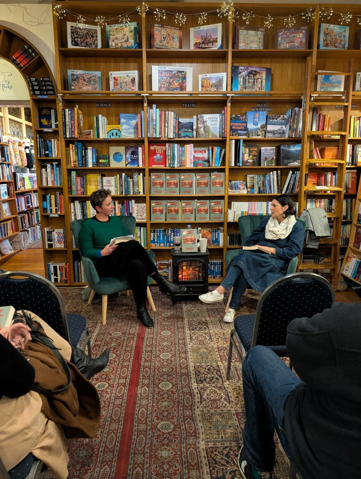 Two women are sitting in green armchairs having a conversation in a bookstore or library. Between them is a small black table with a fireplace, a wine glass, and a cup. The background features wooden bookshelves filled with books and puzzles, and twi
