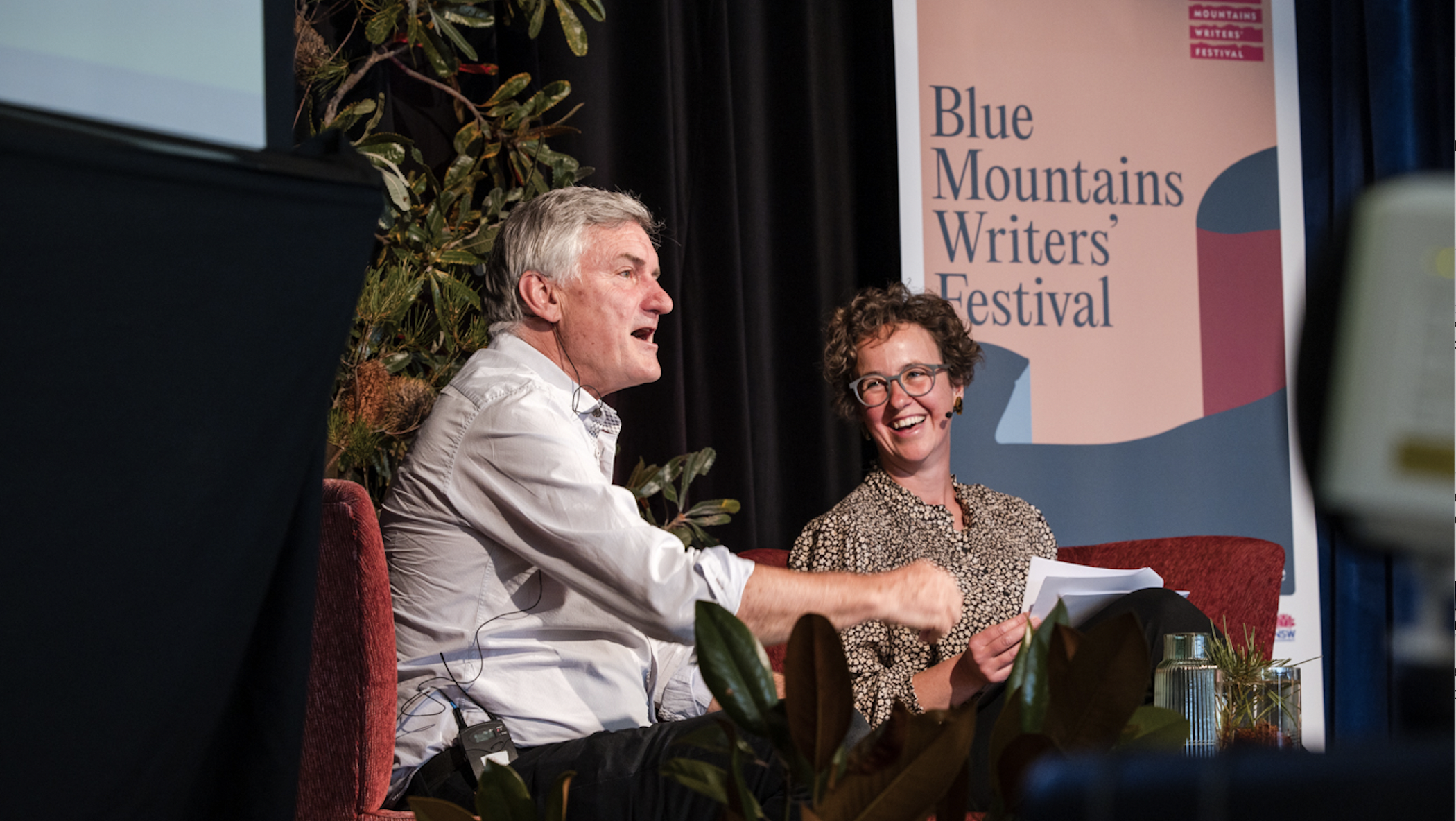 Two people, a man and a woman, sitting on a stage at the Blue Mountains Writers' Festival engaging in a discussion, with a banner and plants behind them.