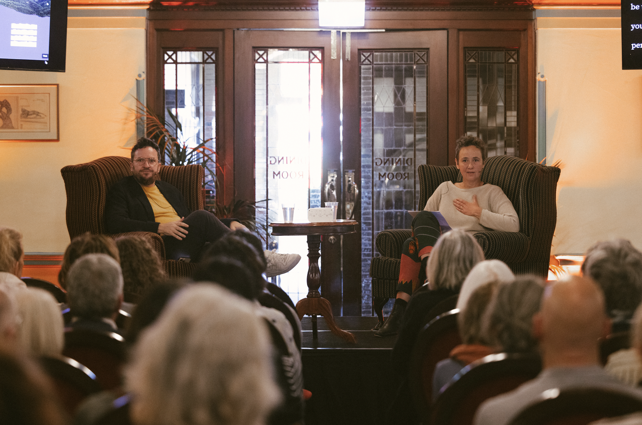Two people seated in armchairs on stage, addressing an audience in a room with wooden doors and windows, a plant, and artwork on the walls.