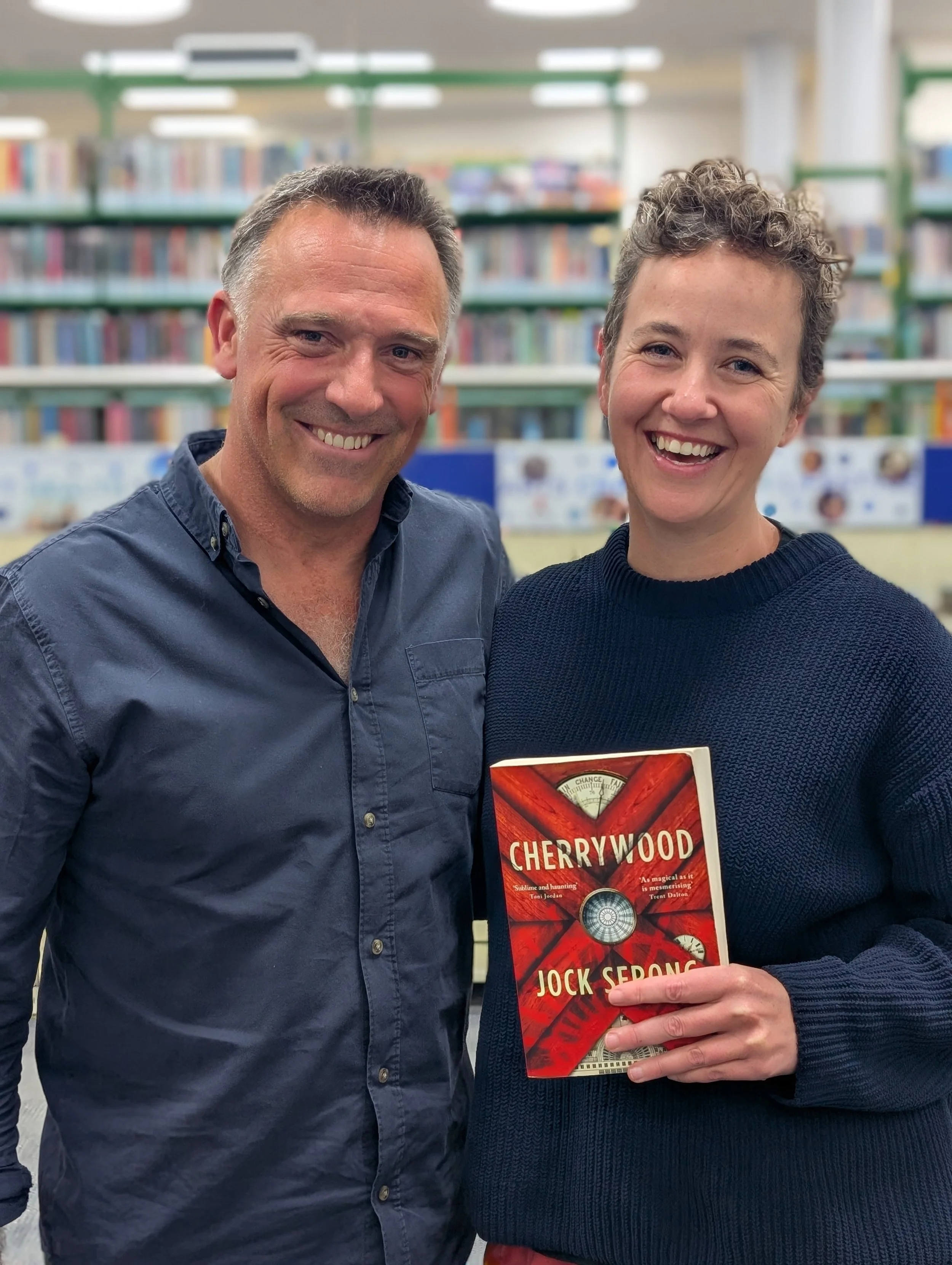 A man and a woman smiling in a bookstore or library, with the woman holding a book titled "Cherrywood" by Jock Serong, and behind them shelves filled with books.