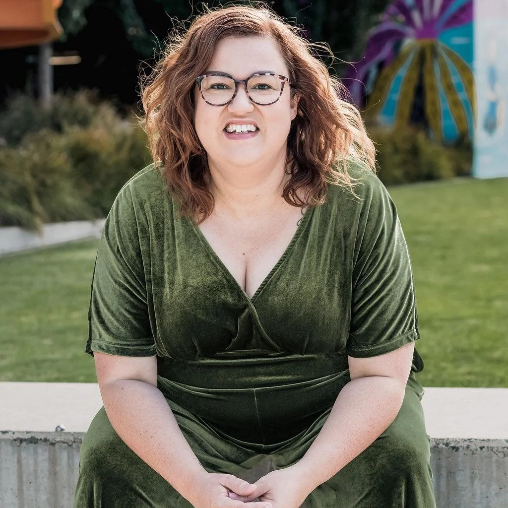 A woman with curly reddish-brown hair, wearing glasses and a green velvet dress, sitting outdoors on a concrete ledge with grass, bushes, and a colorful umbrella in the background.