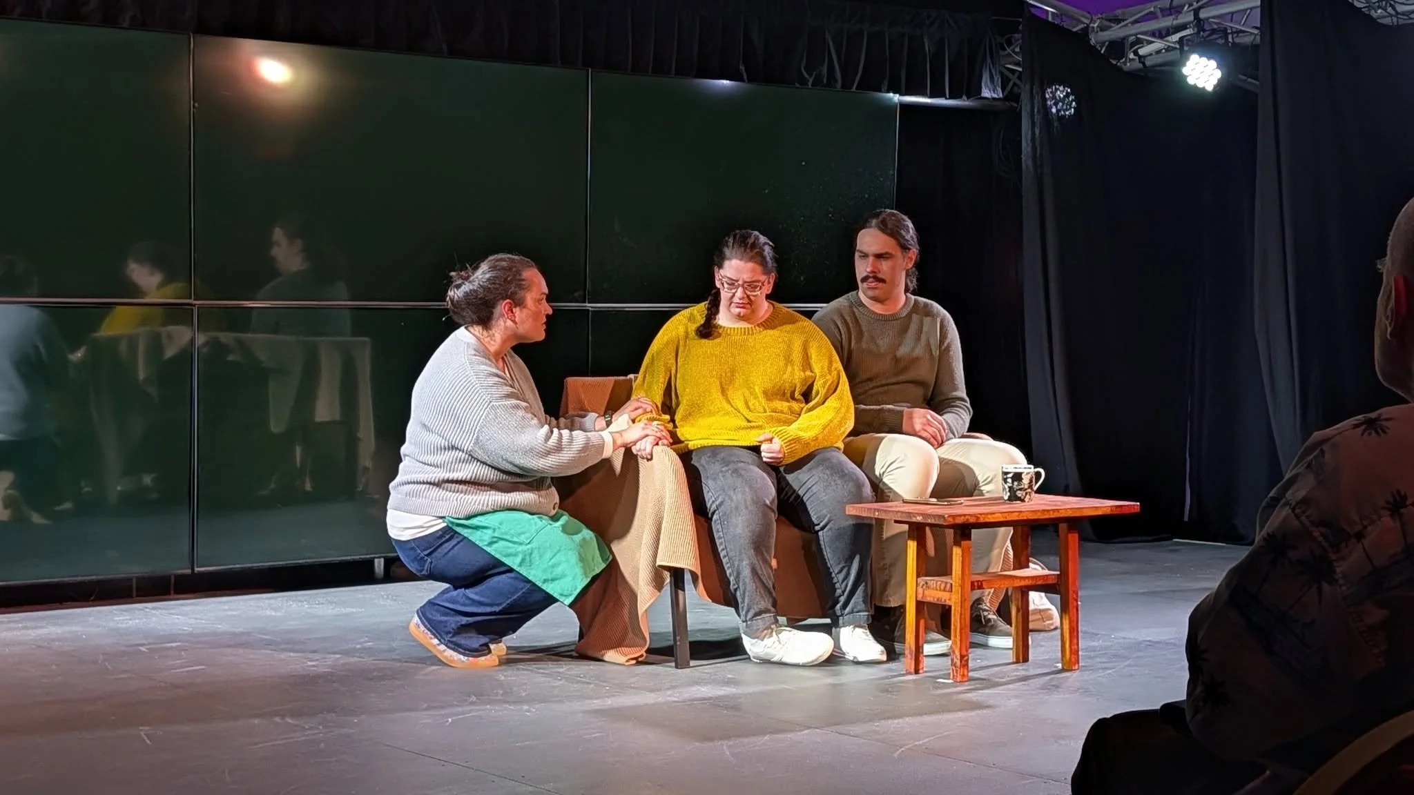 Three people sitting on a couch engaged in a conversation, with one person kneeling and holding hands with the woman in the middle, in a staged setting or theater scene.