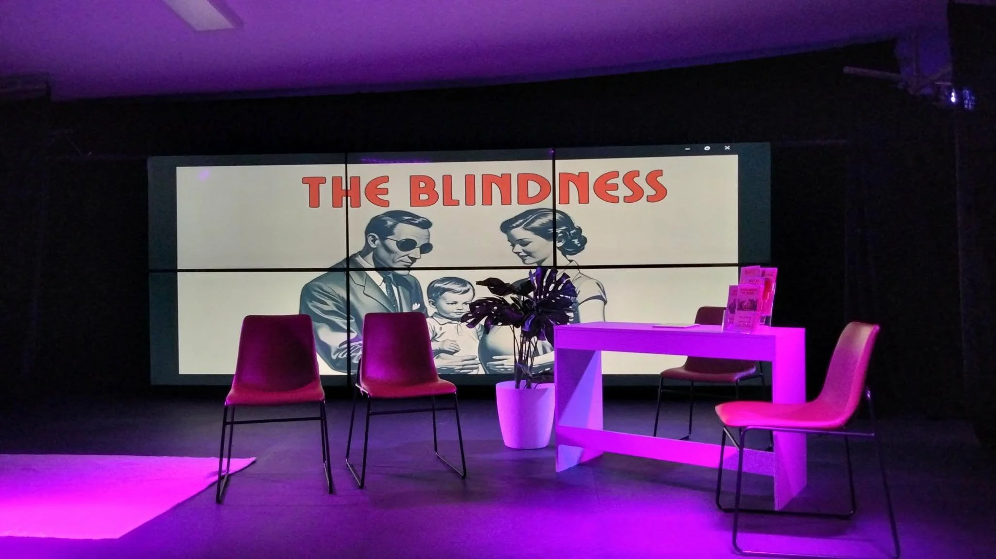A dimly lit room with purple lighting, featuring a large screen displaying a vintage illustration and the words 'The Blindness', surrounded by chairs and a white table.