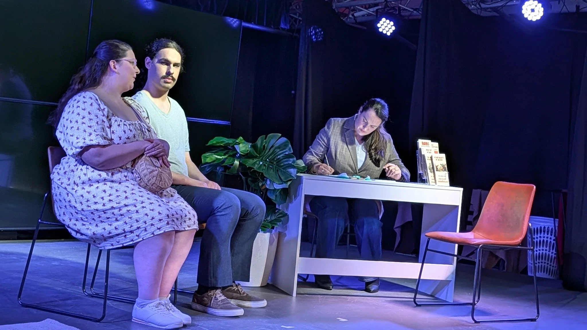 Three people sit on a stage, two women and one man, with a woman working at a desk nearby. There are chairs, a plant, and stage lighting.