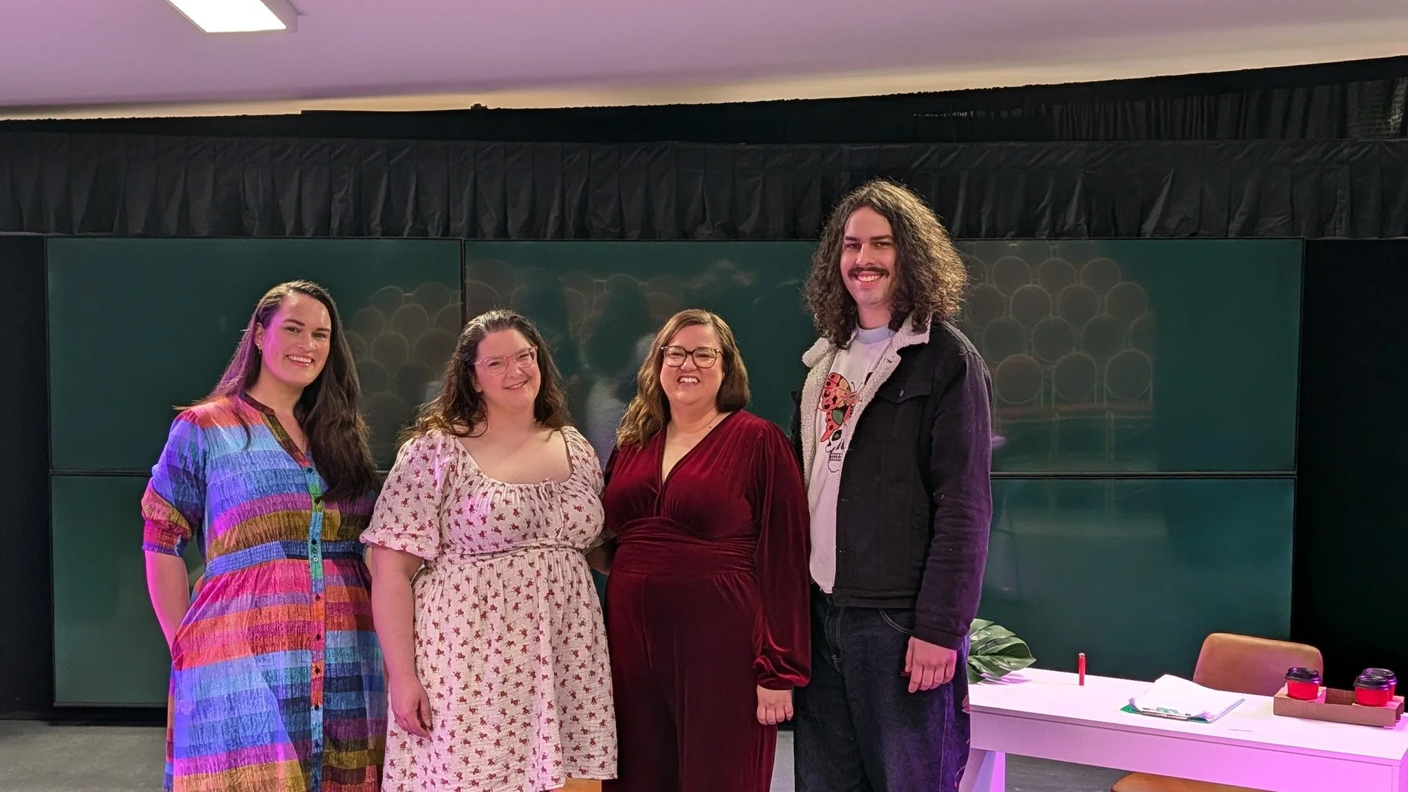 Group of five people standing together indoors, smiling at the camera. Behind them is a dark background with multiple small displays showing or reflecting images.