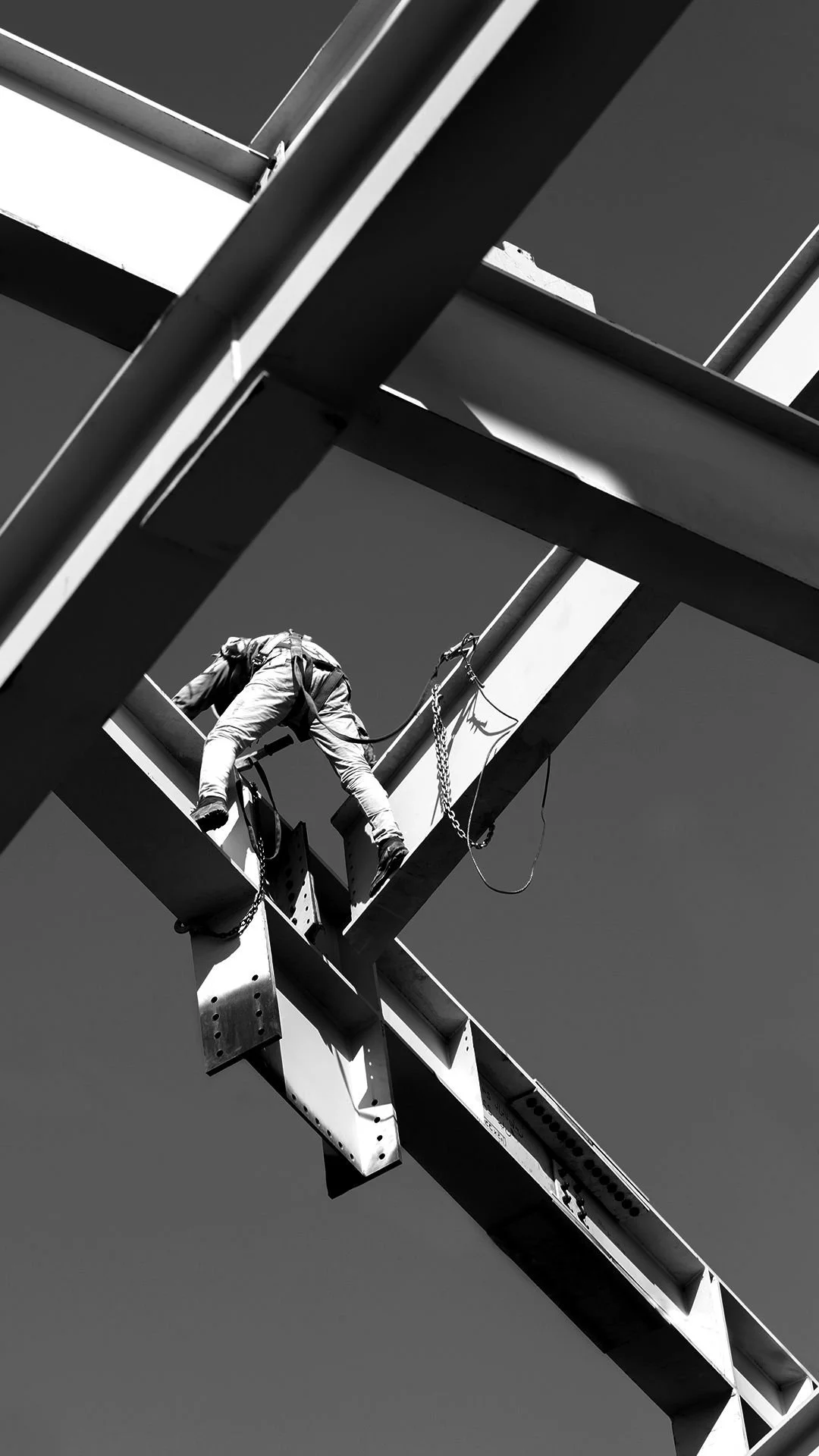 A construction worker on a steel framework, working high above the ground, with a safety harness and chain, against a clear sky.