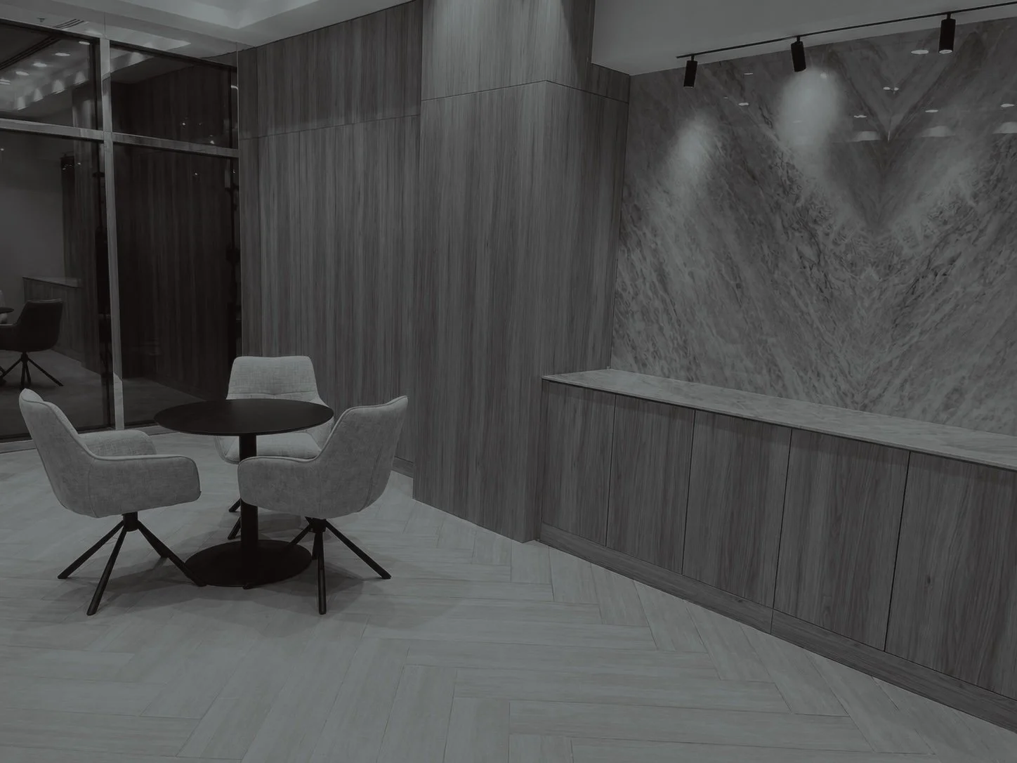 Modern lounge with a round black table and three light gray upholstered chairs, marble wall accents, and track lighting.
