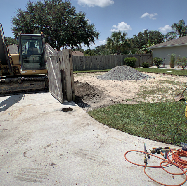 An excavator sits beside a wooden fence next to a gravel pile in a residential yard with grass and an orange hose.