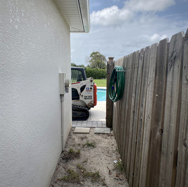 Excavator parked on driveway next to wooden fence and HVAC unit beside a metal-sided house.