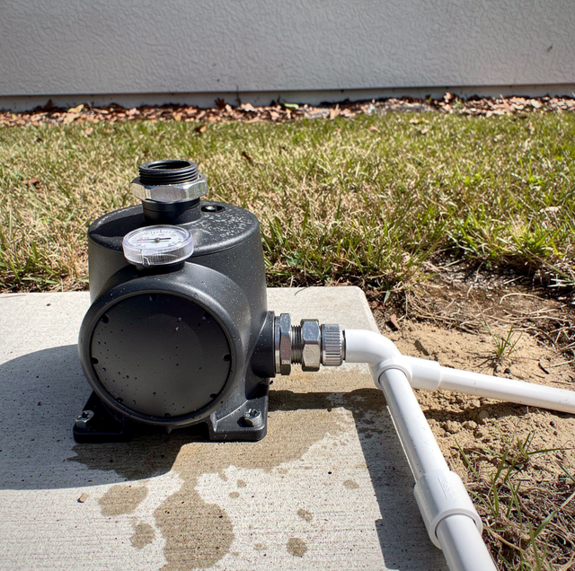 Black pump with gauge mounted on concrete pad next to grass with white pipe and gravel strip.