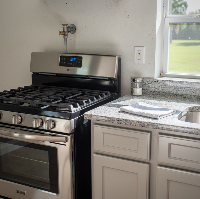 Gas stove with digital display next to granite countertop, cabinets, and window in a kitchen.