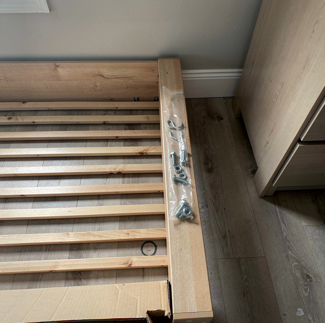 Wooden bed frame with slats and wrapped hardware on flooring next to a wall and door.