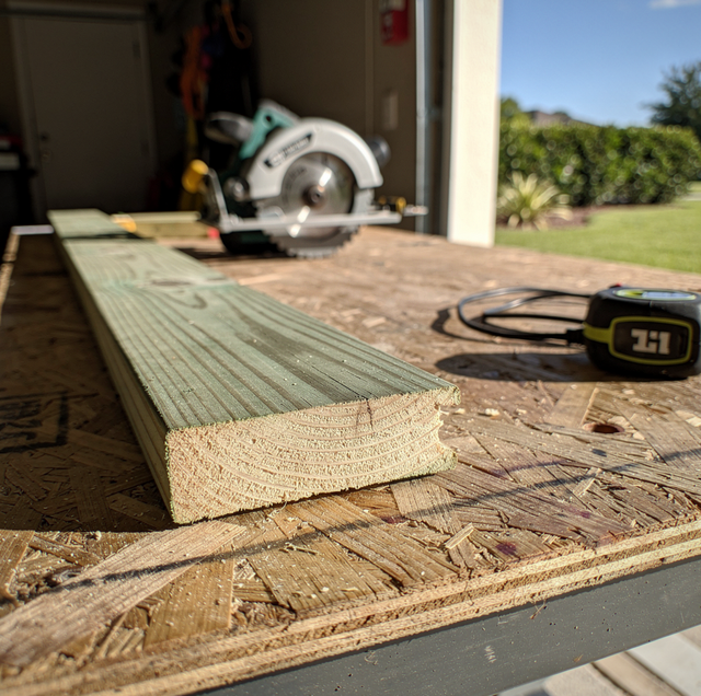 A wooden board and tape measure sit on a work table with a circular saw in a garage, opening to a landscape with trees and gr