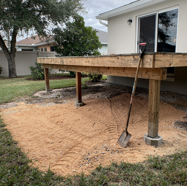 A wooden deck under construction with support posts, a shovel in the dirt beside a white house with large windows.