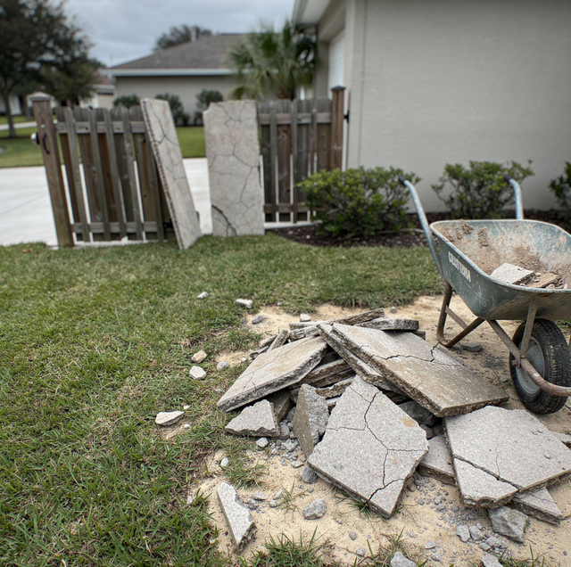 A wheelbarrow and broken concrete slabs sit on grass next to a wooden fence and white house.