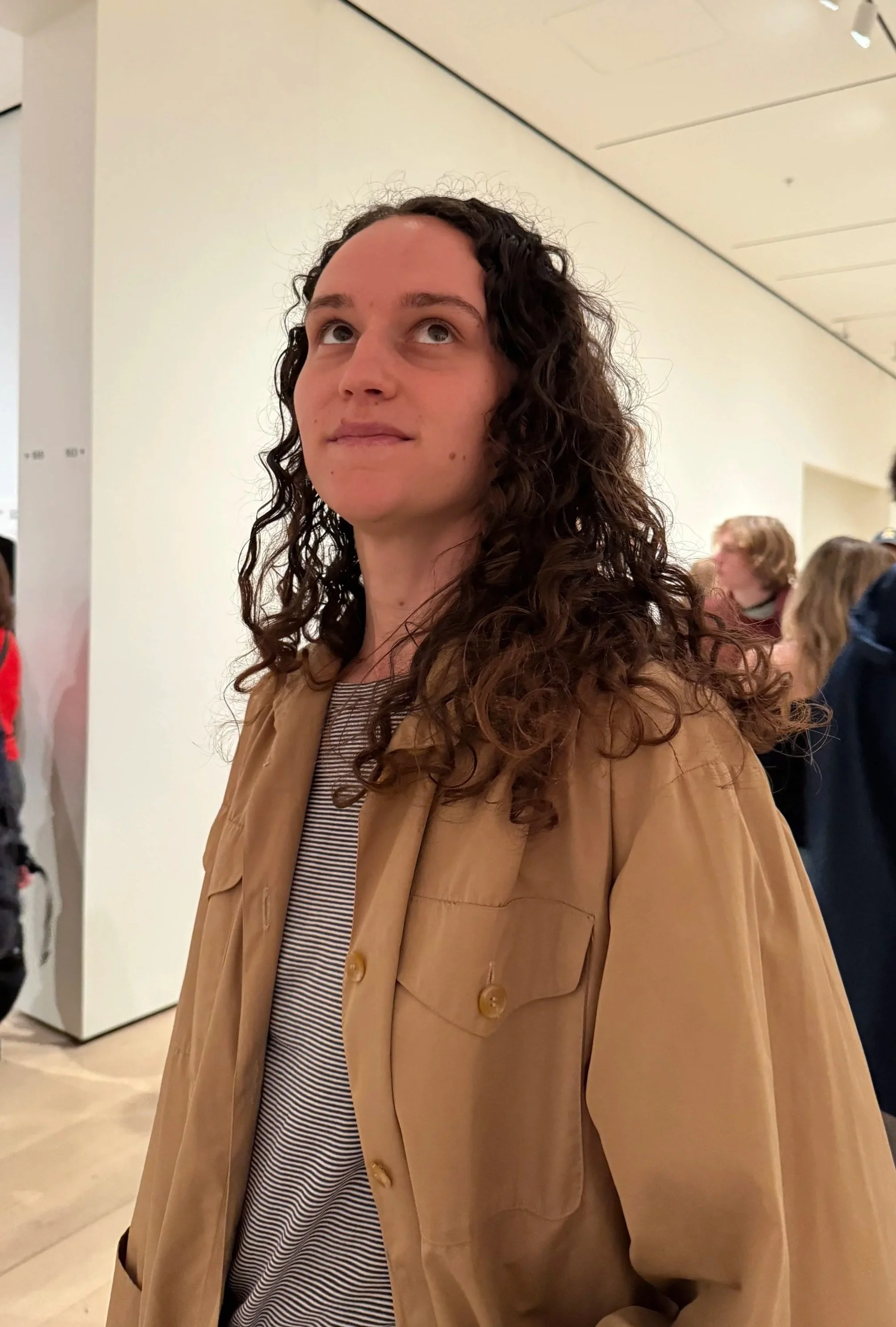 A woman with curly dark hair wearing a tan jacket and striped shirt, standing in a gallery or museum.