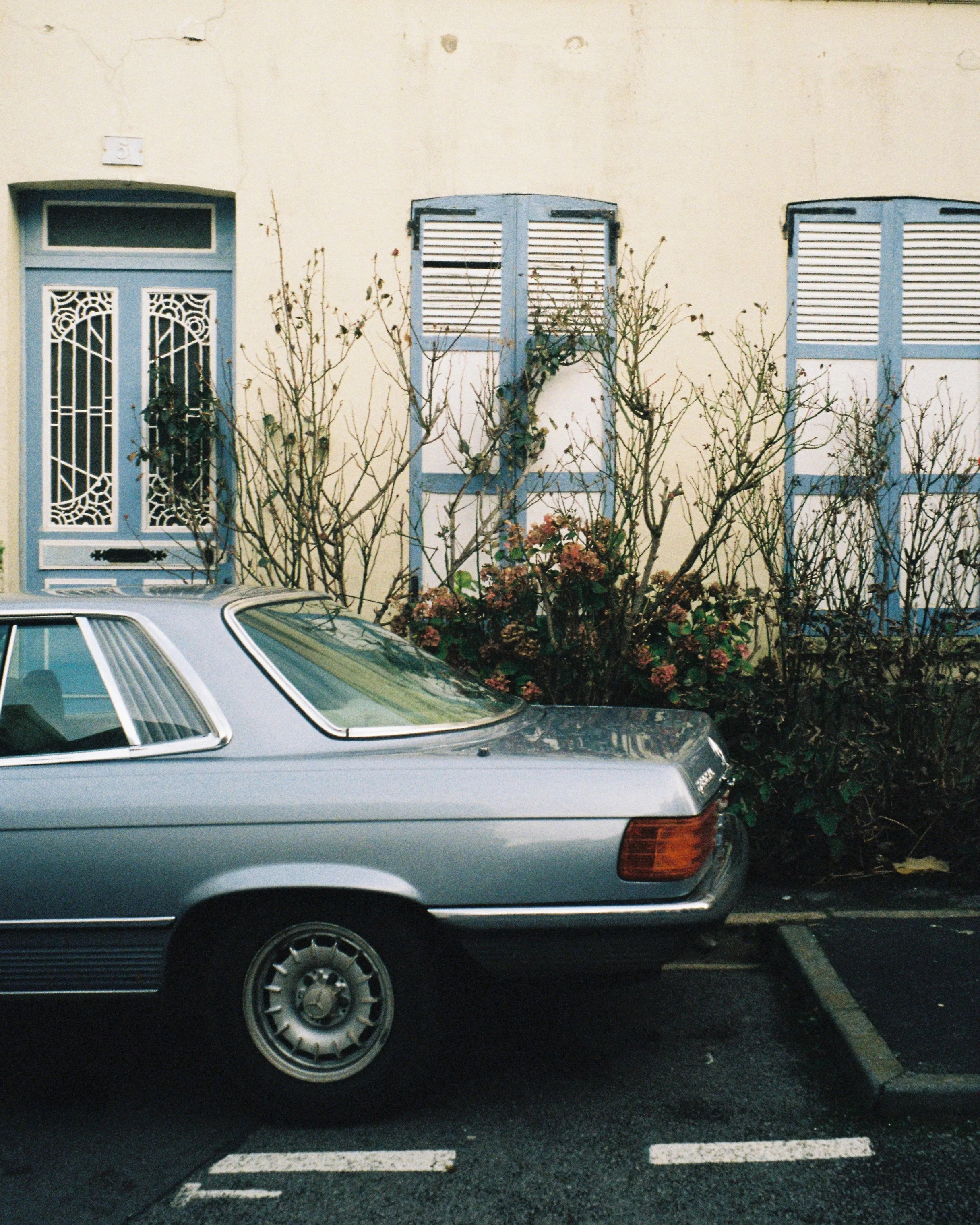 A vintage car parked in a parking lot, with part of the car's tail and tail light visible, and a background of a beige building in Étretat with closed window shutters and a small tree or bush in front.