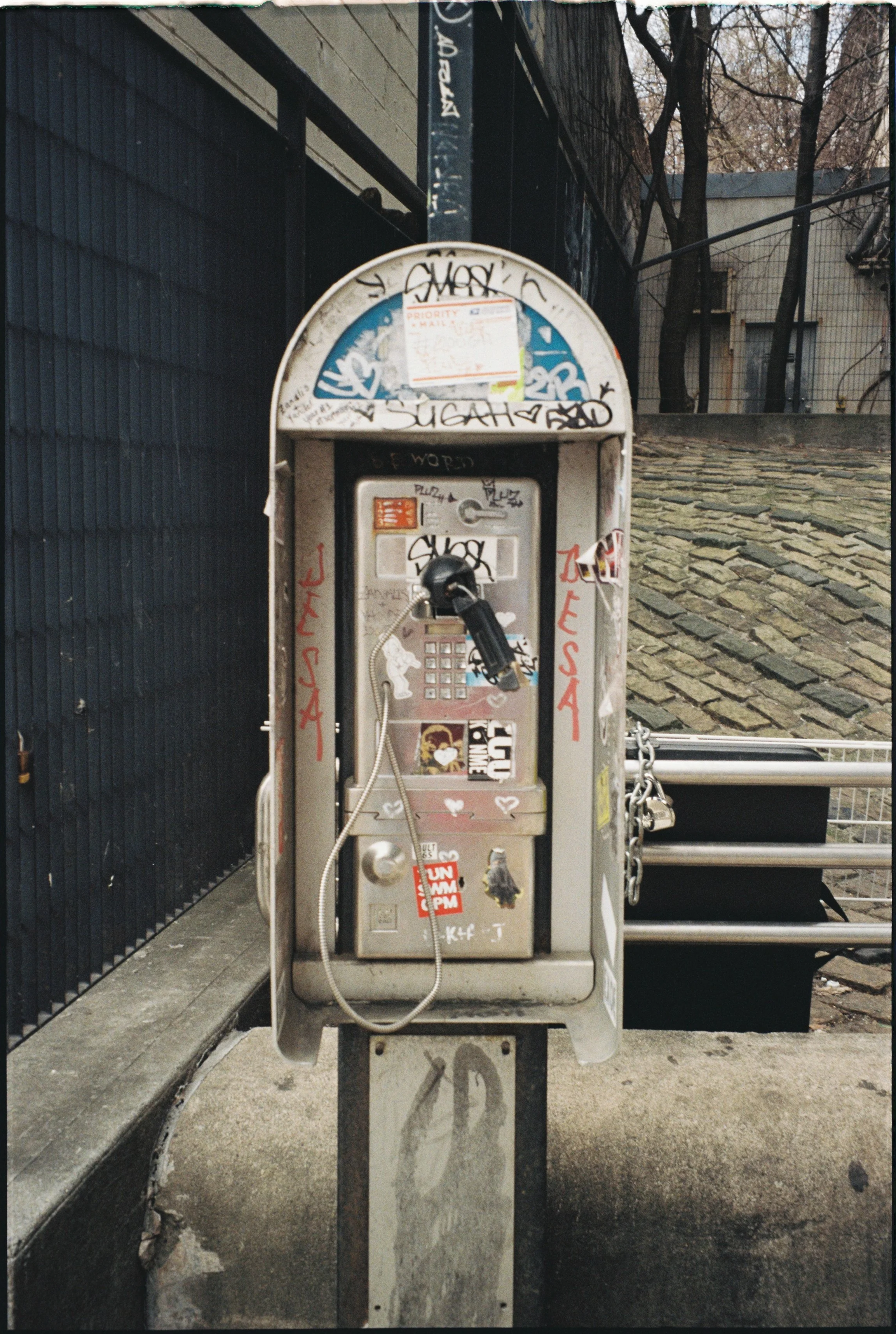 A public payphone covered in graffiti and stickers, situated on a sidewalk next to a black metal fence and a stone wall with a chain barrier. Background includes trees and a building.
