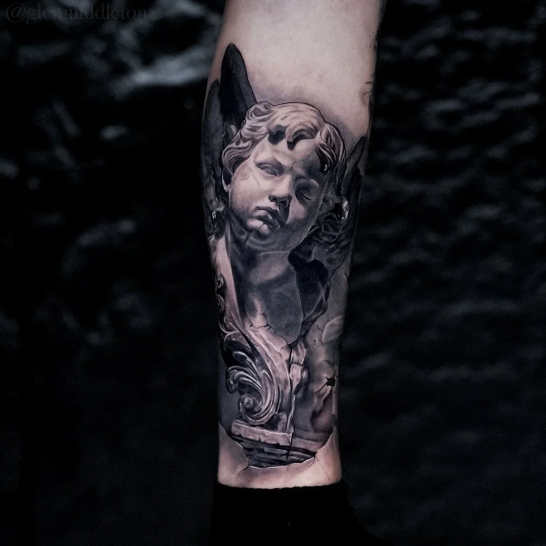 Black and grey forearm tattoo by Glen Middleton featuring a marble cherub sculpture with baroque ornamental detailing.