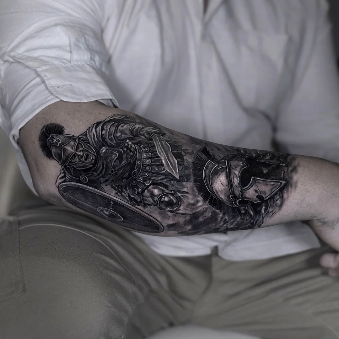 Black and gray tattoo of a Roman soldier on a person's forearm, featuring a helmet, shield, and spear.