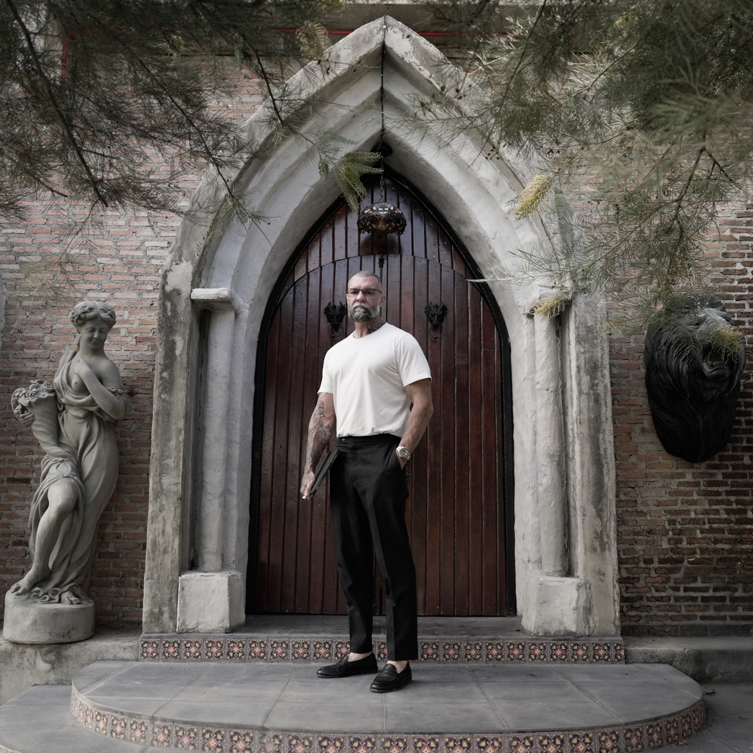 Portrait of sculptural tattoo artist Glen Middleton standing before a Gothic stone archway, reflecting the classical architecture and fine art influences behind his large scale black and grey tattoo work.