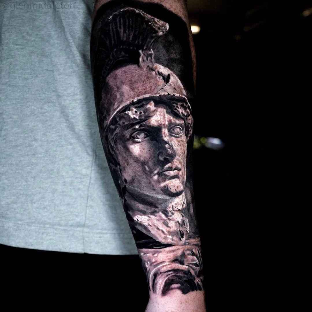 Black and grey forearm tattoo by Glen Middleton featuring a Roman soldier with shield and helmet in sculptural realism.