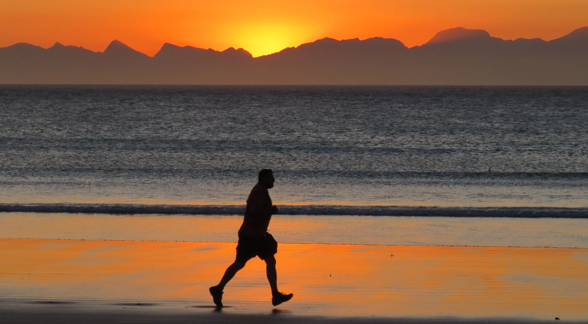 A person jogging on the beach during sunset with mountains in the background.
