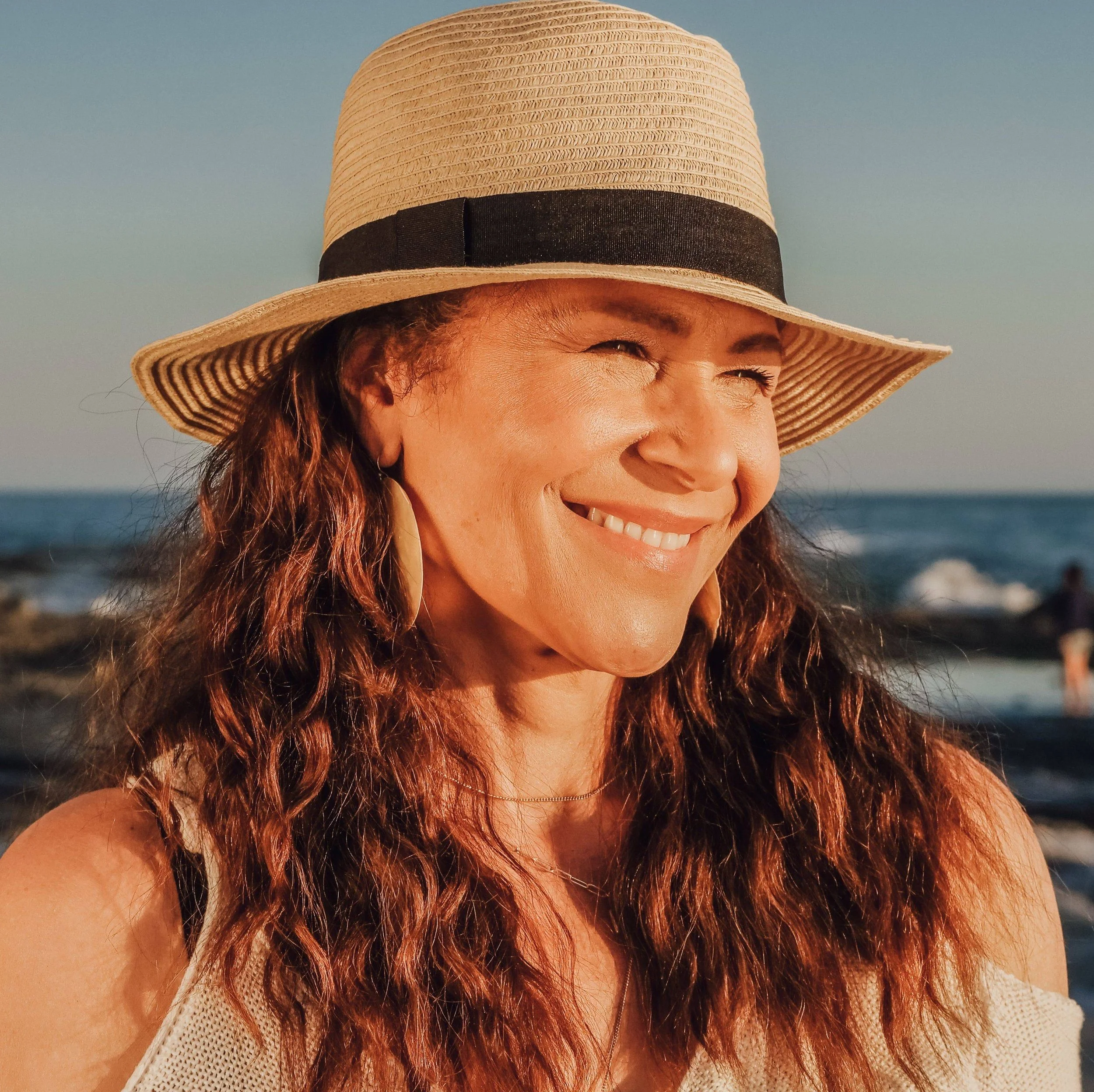 Close-up of a smiling woman wearing a wide-brimmed straw hat with a black ribbon, with her face partially visible, against a background of a clear sky and ocean.