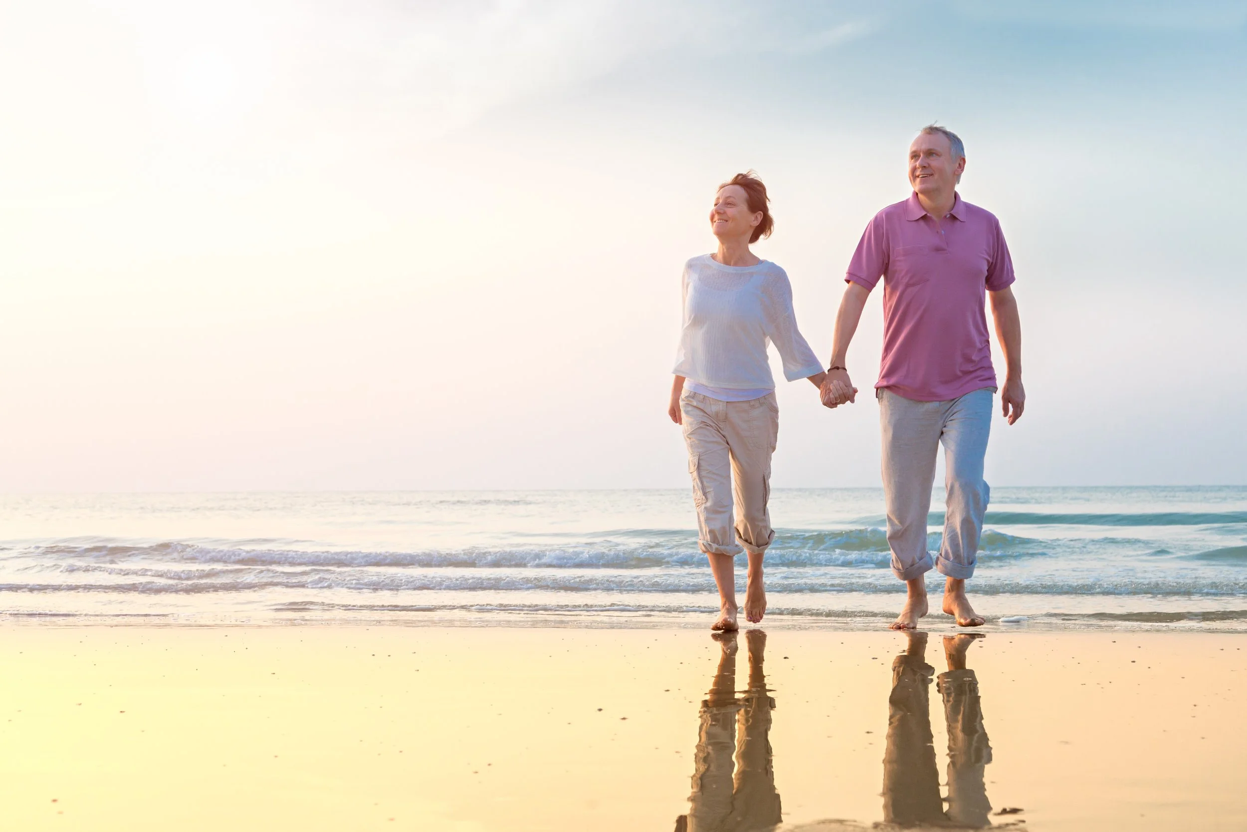 A happy elderly couple walking hand in hand along the beach during sunset, reflected in the shallow water.