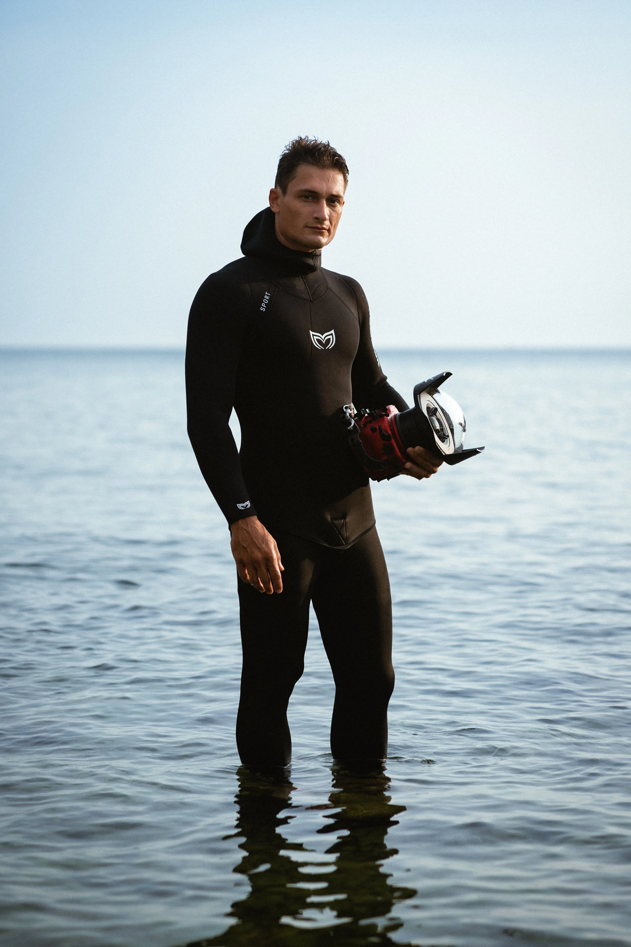 Man standing in water at the beach, holding a helmet, dressed in black sportswear, with a calm sea and sky in the background.